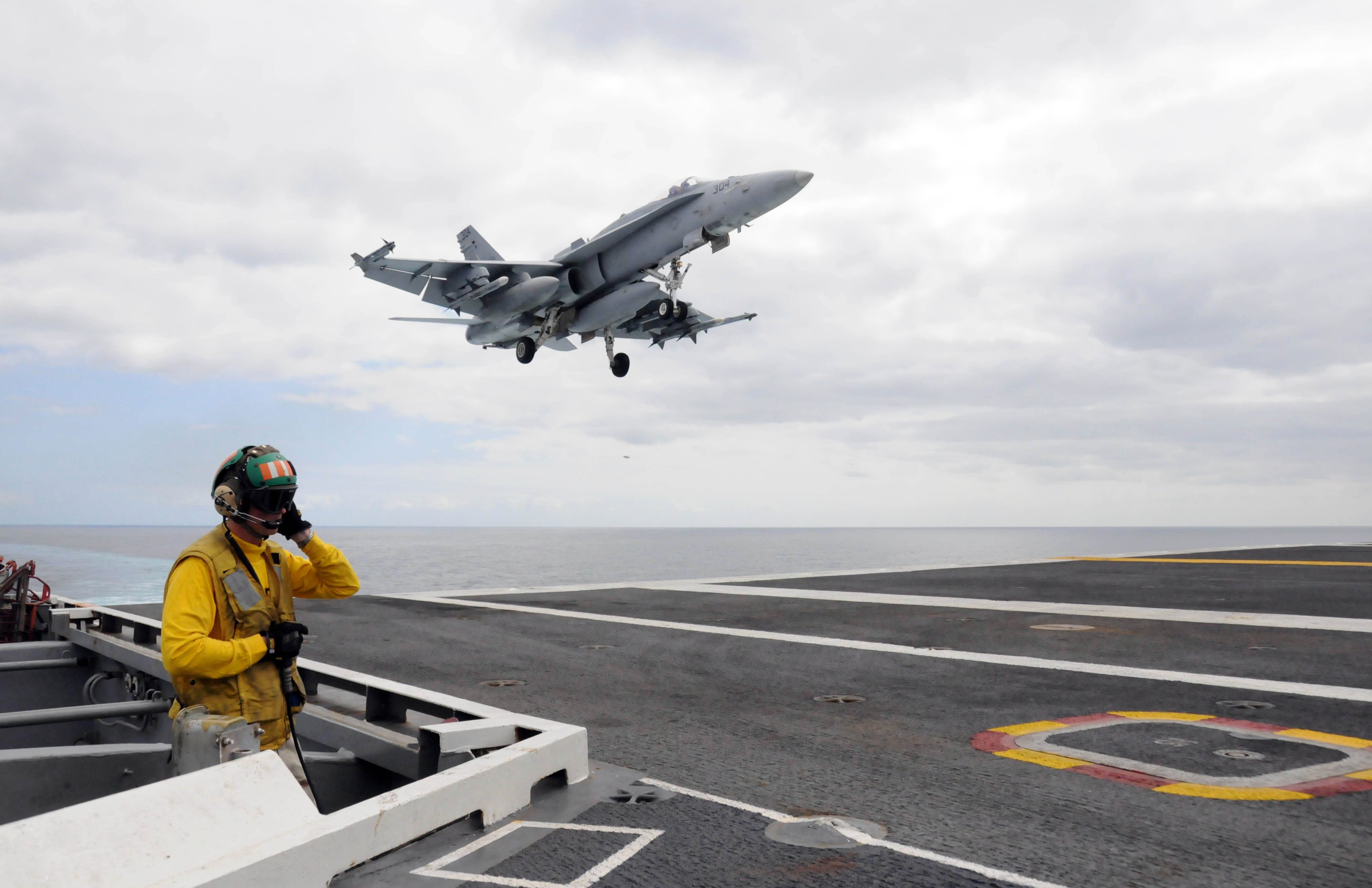     Of A Soldier In Yellow Watching A Jet Land On An Aircraft Carrier