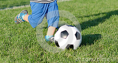     The Legs Of A Little Boy Kicking A Soccer Ball On A Green Sportsfield