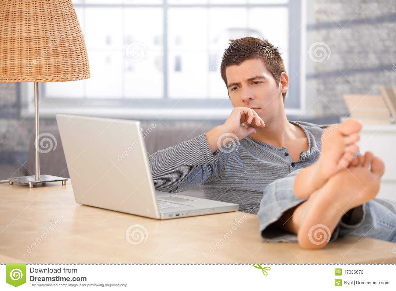 Young Man Sitting At Home With Bare Feet On Desk Thinking Looking At