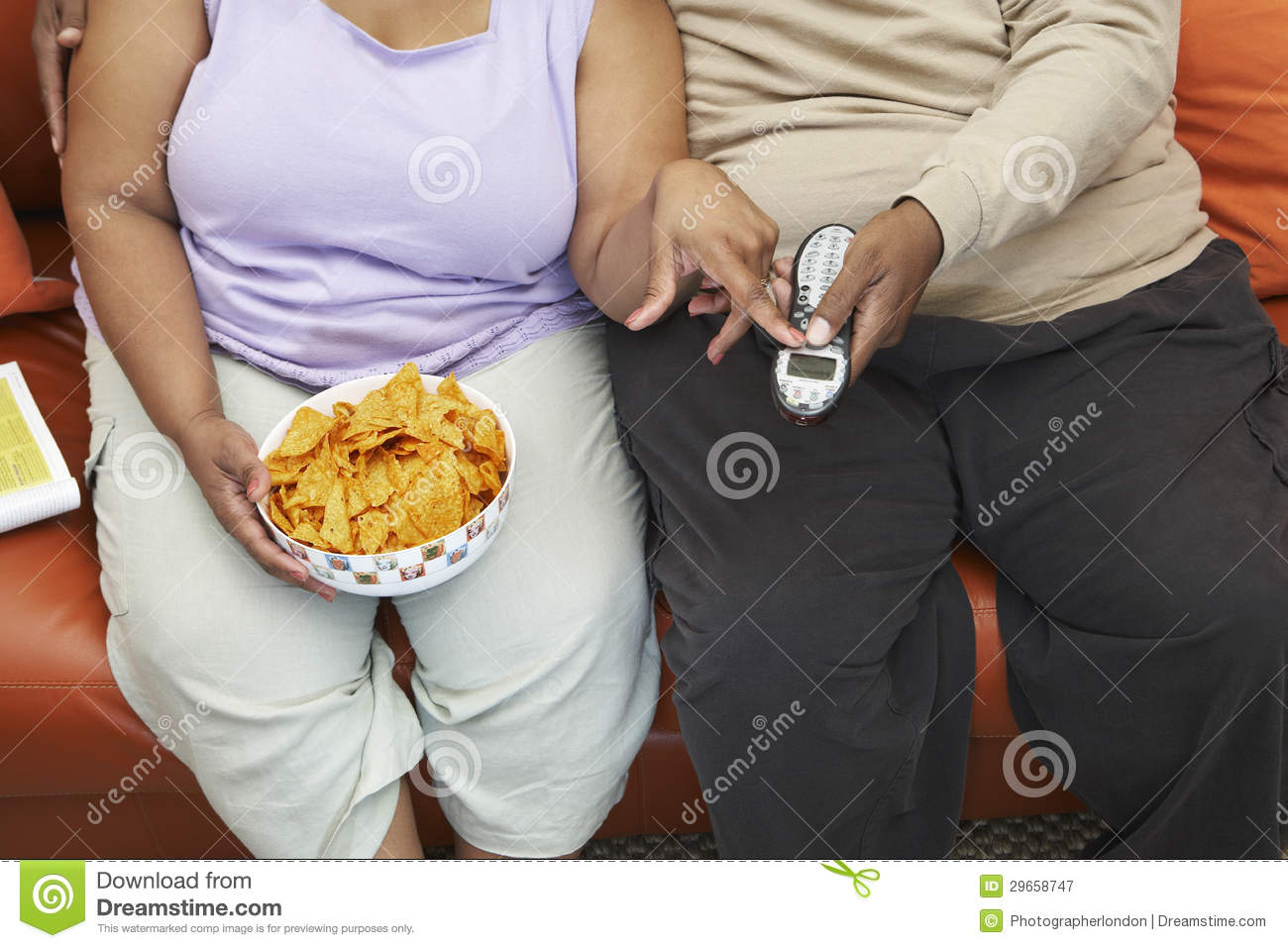 Midsection Of An Obese Couple Sitting On Couch With Nachos And Remote    