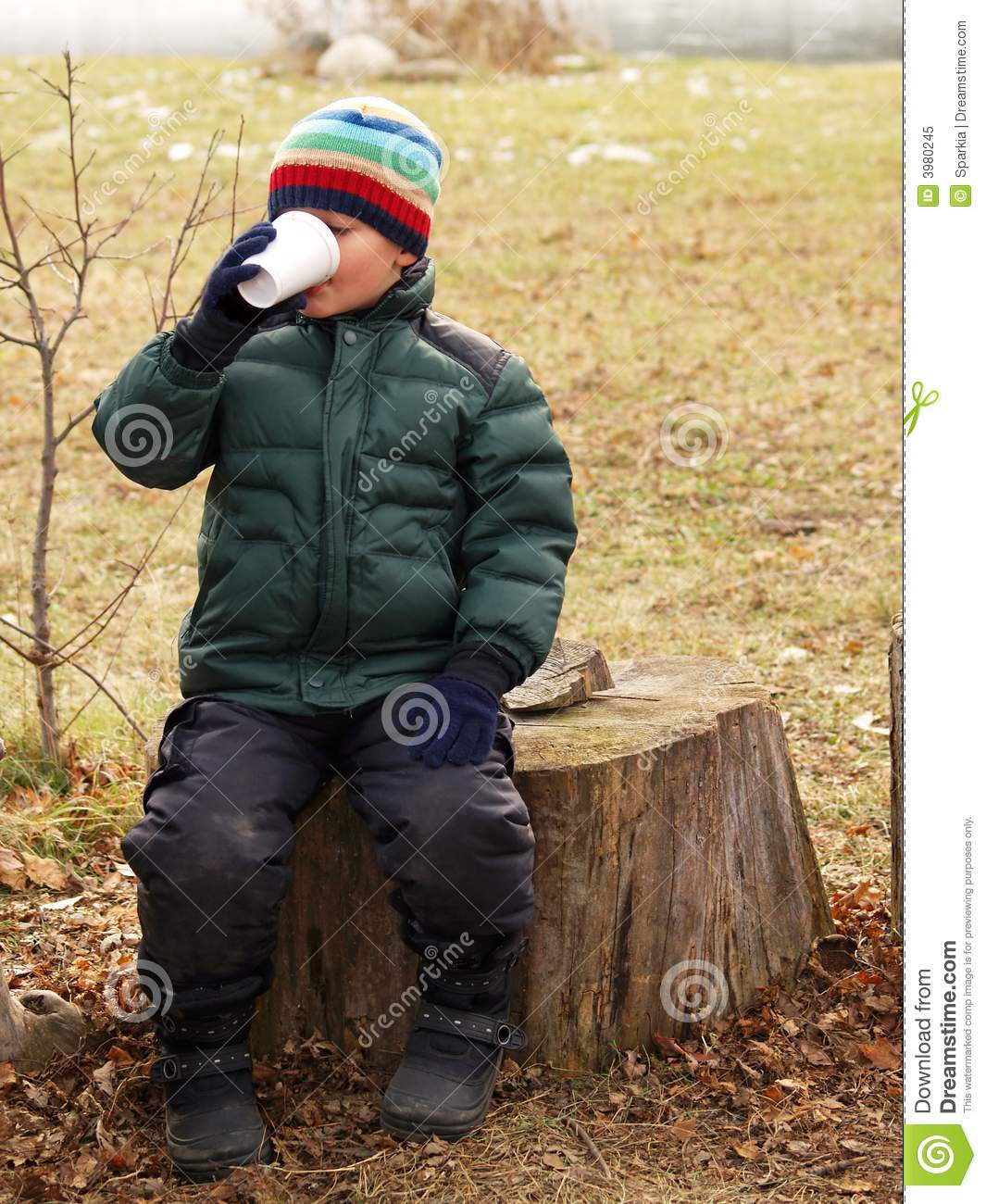 Young Boy Drinking Hot Chocolate While Sitting On A Tree Stump Outside    