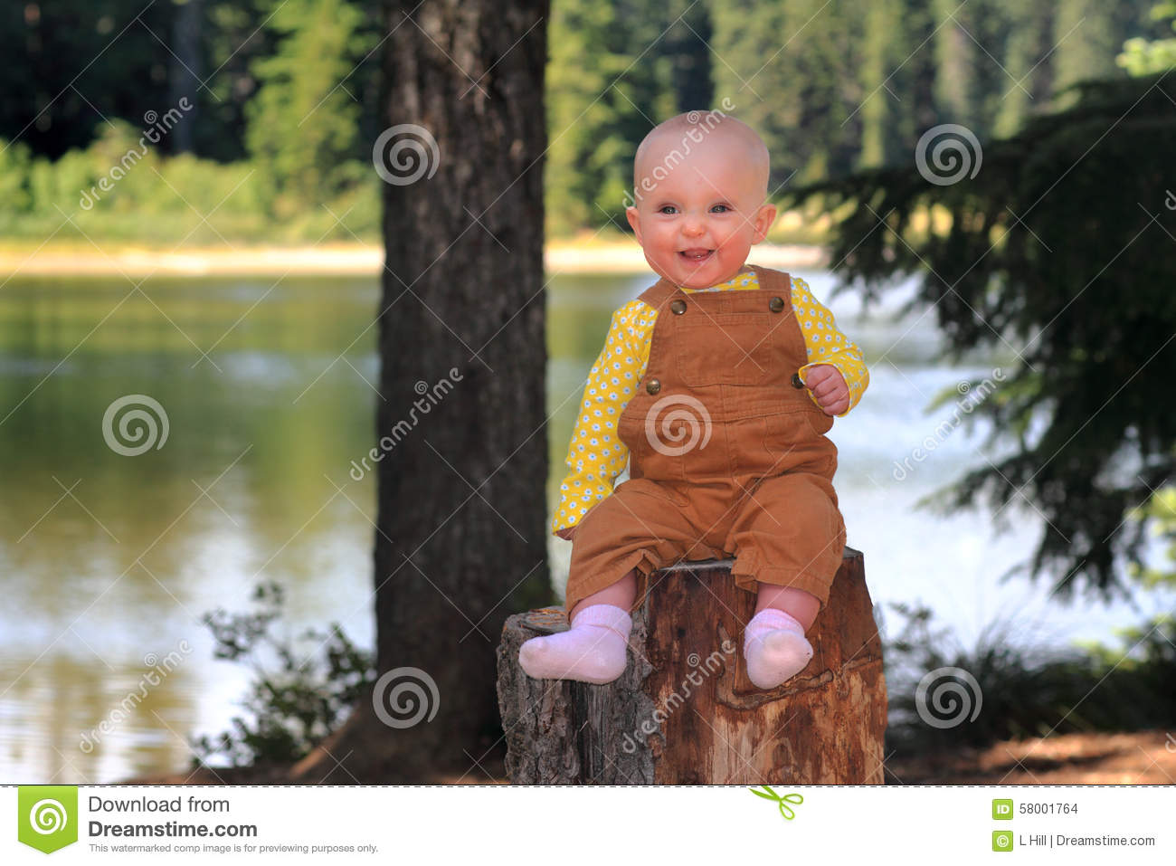 Cute Smiling Baby Wearing Overalls Sitting On A Stump In Front Of A