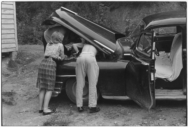 Two Men Working Under Car Hood  Young Woman Leaning On Fender Watching    